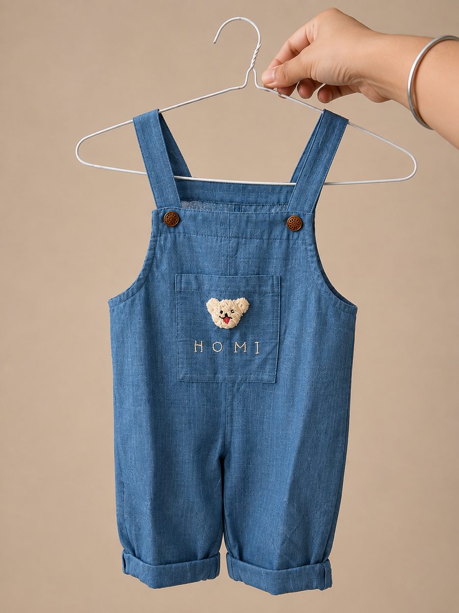 Child's blue cotton dungarees on a hanger, with an embroidered name and a teddy on the pocket